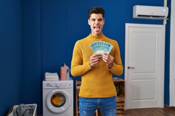 Sticker - Young hispanic man at laundry room holding brazilian reals sticking tongue out happy with funny expression.