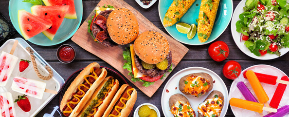 Wall Mural - Summer BBQ food table scene. Hamburgers, hot dogs, potatoes, corn and cold treats. Top view over a dark wood banner background.