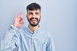 © Krakenimages.com - Young hispanic man with beard standing over blue background smiling positive doing ok sign with hand and fingers. successful expression.