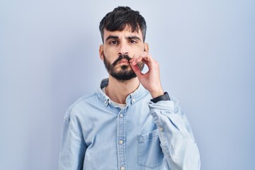 Poster - Young hispanic man with beard standing over blue background mouth and lips shut as zip with fingers. secret and silent, taboo talking