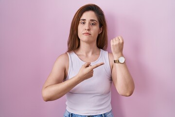 Poster - Brunette woman standing over pink background in hurry pointing to watch time, impatience, looking at the camera with relaxed expression