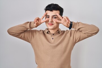 Poster - Non binary person standing over isolated background doing peace symbol with fingers over face, smiling cheerful showing victory