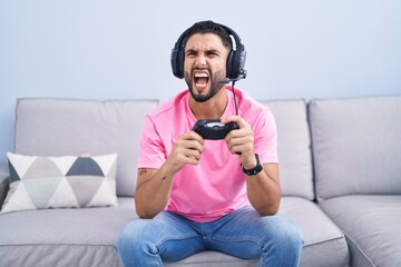 Canvas Print - Hispanic young man playing video game holding controller sitting on the sofa angry and mad screaming frustrated and furious, shouting with anger. rage and aggressive concept.