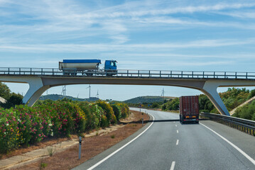 Canvas Print - Container truck driving on a highway while another dump truck passes over a bridge.