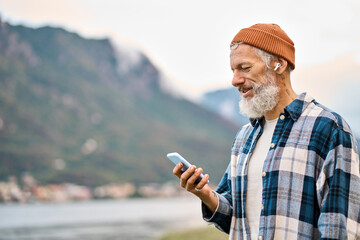 Cool bearded healthy older elder hipster man standing in nature park wearing earbud using mobile phone. Mature traveler enjoying freedom, active leisure lifestyle, listening music on smartphone.