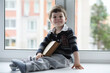 © alexkich - The little boy is reading a book. The child sits at the window and prepares for lessons. A boy with a book in his hands is sitting on the windowsill.