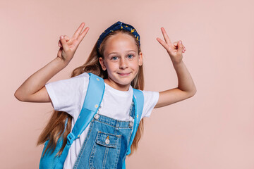 Blond school girl with blue eyes, wears a blue backpack and denim overalls, does peace victory sign with two hands posing at studio isolated over beige background. Back to school concept.