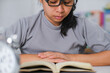 © Pornpimon - Closeup image of Asian woman sitting at a table reading a book in the living room.