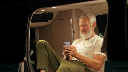 Wall Mural - Older man sitting inside rv camper van with tent on it on vacation using mobile phone. Senior mature traveler holding smartphone enjoying free internet or checking rentals in camping at night.