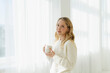 © Westend61 - Blond woman holding coffee cup by translucent curtain