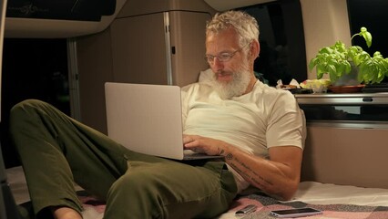 Wall Mural - Older man sitting in rv camper van using laptop at night. Mature active traveler digital nomad holding computer on lap remote working online on wireless technology device.