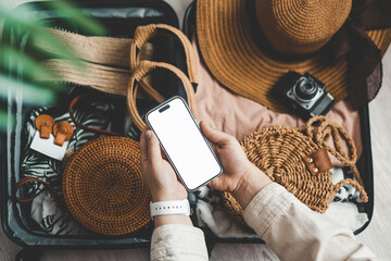 Top view of female hands holding smartphone with blank screen near suitcase with beach accessories on wooden floor