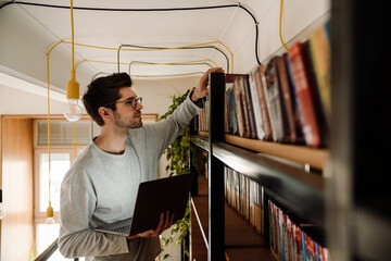 Man student using laptop and looking for book while standing in library