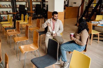 Wall Mural - Indian man talking with сollege classmate and drinking coffee while sitting in library