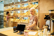 © pressmaster - Mature female clerk or owner of bakery entering data in computer while counting total cost of purchase or upgrading prices for bread