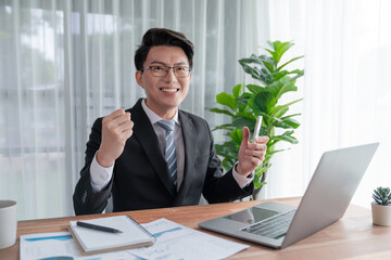 Excited Asian businessman celebrates success at office desk. Happy office worker achieves business goals with data analysis or marketing planning. Business winner celebratory expression. Jubilant