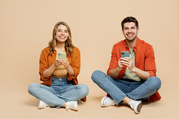 Wall Mural - Full body fun young couple two friends family man woman wearing casual clothes together sitting hold in hand use show mobile cell phone isolated on pastel plain beige color background studio portrait.