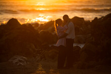 A Young Couple In Love Stands On A Rocky Ocean Shore During A Golden Sunset.