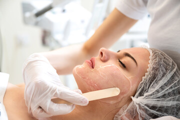 Wall Mural - Close up of beautician hands with wooden spatula applying cosmetic product gel peeling mask to beautiful young woman face. Skincare and cosmetology spa concept.