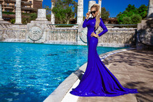 Elegant Luxury Evening Fashion. Glamour, Stylish Elegant Woman In Blue Long Evening Gown Dress Is Posing Nead The Pool In Luxury Hotel Outdoor. Female Model In Amazing Long Dress.