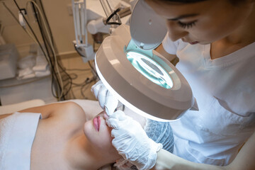 Wall Mural - Beautician squeezing acne and pimples from young woman's nose using lamp and magnifying glass in beauty spa clinic.