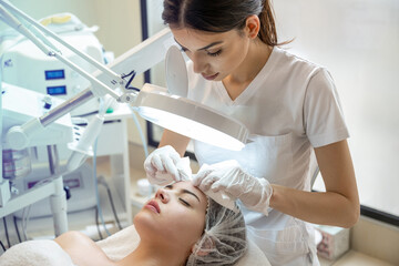 Wall Mural - Beautician squeezing acne and pimples from young woman's nose using lamp and magnifying glass in beauty spa clinic.
