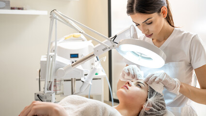 Wall Mural - Beautician squeezing acne and pimples from young woman's nose using lamp and magnifying glass in beauty spa clinic.
