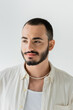 © LIGHTFIELD STUDIOS - Portrait of young bearded homosexual man in beige shirt, sleeveless t-shirt and pearl necklace looking away while standing isolated on grey