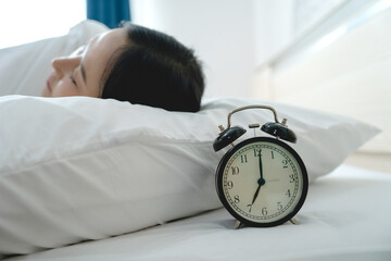 Alarm clock that is going to ring at seven in morning on background of woman sleeping in bed.