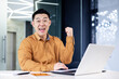 © Liubomir - Successful asian businessman celebrating victory success, portrait of man in office in casual shirt, employee smiling and looking at camera satisfied with achievement holding hand up victory gesture