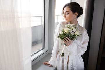 young woman with engagement ring on finger standing in white silk robe and holding bridal bouquet ne