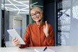 © Liubomir - A smiling young woman in a headset sitting in the office, holding a tablet in her hand and greeting clients and partners via video call.