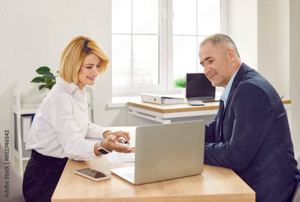 Smiling woman consultant and client sitting at desk using laptop discussing insurance banking services in office. Mature man having consultation with financial advisor, real estate, insurance agent