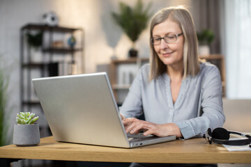 Wall Mural - Smiling middle-aged woman in uncollared blouse typing on portable computer while sitting at writing desk in home office. Attractive business lady in glasses working remotely using electronic device.