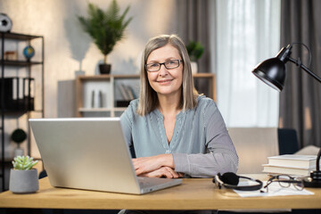 Portrait of happy mature lady in everyday clothes posing with laptop on writing desk in cozy workplace indoors. Cheerful caucasian female in spectacles using remote office for managing business.