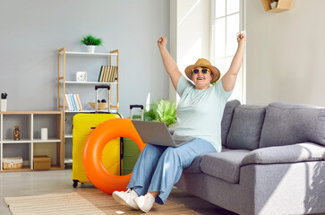 Funny female tourist books cheap airline ticket. Happy fat woman in summer hat and sunglasses sitting on sofa with laptop at home in room with suitcases and beach ring. Holiday vacation travel concept