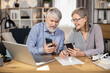 © Sofiia Shunkina - Close up view of mature businesspeople with cell phones looking at each other while staying in comfortable workplace. Senior husband and wife running business together using modern technologies.
