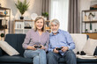 © sofiko14 - Focused male and female adults around 60 using multiple buttons on game pads during home entertainment. Cheerful couple of mature people improving teamwork skills while playing games together.