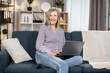 © Sofiia Shunkina - Caucasian woman of 60s resting on soft couch with portable laptop and browsing social media. Smiling female person dressed in casual wear using modern gadgets at cozy home.
