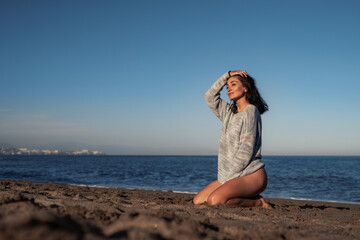 Wall Mural - beautiful brunette woman in a sweater and swimsuit sits on the sand by the sea enjoys the environment