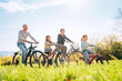 © Soloviova Liudmyla - Smiling father and mother with two daughters during summer outdoor bicycle riding. They enjoy togetherness on green high grass meadow. Happy parenthood and childhood or active sport life concept image