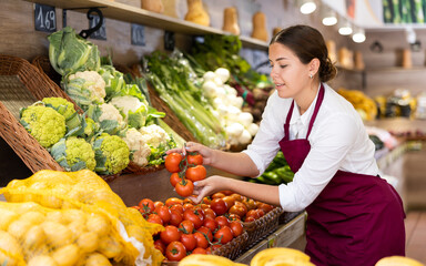 Delighted young saleswoman placing fresh tomatoes on food stall in grocery store