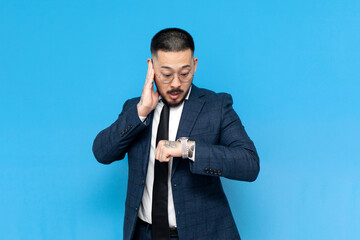 Wall Mural - successful asian businessman in suit and glasses is late and looks at his watch on blue isolated background