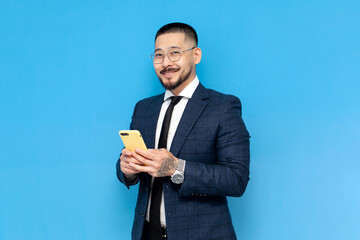 Wall Mural - successful asian businessman in suit and glasses uses smartphone on blue isolated background and smiles