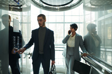 Poster - Young businessman pressing button in elevator while standing against female colleague with mobile phone by ear talking to client