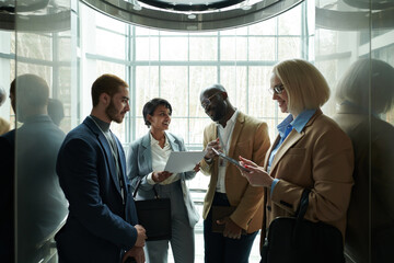 Poster - Group of successful intercultural delegates in formalwear preparing reports for conference while standing in elevator and moving upwards