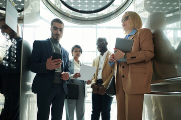 Poster - Group of intercultural entrepreneurs revising points of their reports in smartphone and on paper while standing in moving elevator
