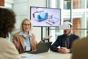 Leinwandbilder - Confident mature business coach explaining something to employees at working meeting while making presentation of new vr headset