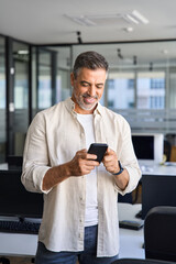 Wall Mural - Vertical portrait of middle aged Latin or Indian business man using phone mobile technology application for online work communication. Smiling mature Hispanic businessman holding smartphone in office.