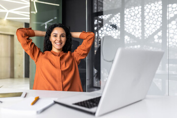 Wall Mural - Portrait of successful Hispanic businesswoman, female worker at workplace resting after successful completion of work, woman smiling and looking at camera, satisfied with herself, hands behind head.